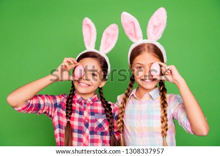 Close up photo of two pretty little age girls holiday concept with bunny ears on head hiding one eye behind easter colored eggs wearing casual checkered plaid shirts isolated on green background