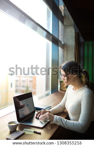 Young hipster girl in fashionable glasses experienced creative designer working on pc laptop computer, sitting in modern co-working space. Female online booking via notebook device 