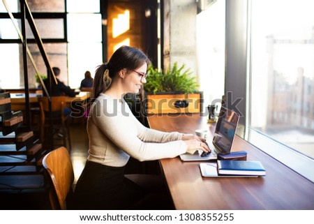 Happy smiling hipster girl skilled social media content writer freelance working on laptop computer while sitting in co-working coffee shop.Female blogger typing article on website via notebook device