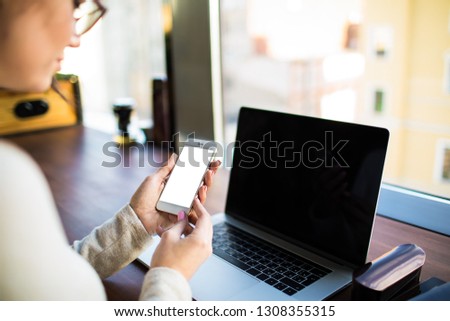 Female web editor holding cell telephone with blank mockup display background for promotional content during distance work on laptop computer, sitting at desktop in modern office interior
