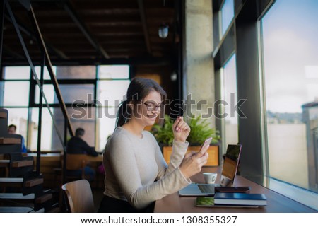 Happy smiling woman in casual wear and fashionable spectacles prosperous freelance worker watching video on smartphone during webinar on laptop computer, sitting in co-working space