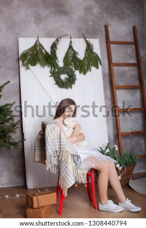 Cute little blonde girl in white dress sits in an embrace with bear. child opens gift Christmas eve. cozy house. Eco-style room. Christmas atmosphere, comfort. Dreamy girl. New Year. Merry Christmas