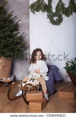 Little curly girl sits in sleigh and holds gift in her hands. child opens gifts at home. Wooden sleigh. cozy house. child receives Christmas present in living room. Merry Christmas. New Year 2020