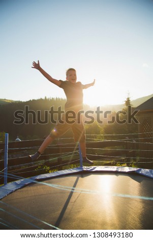 Cute funny cheerful young kid jumping outdoors at trampoline at countryside in soft sunset sunlight. Healthy lifestyle and happy summer vacations concept. Vertical color photography.