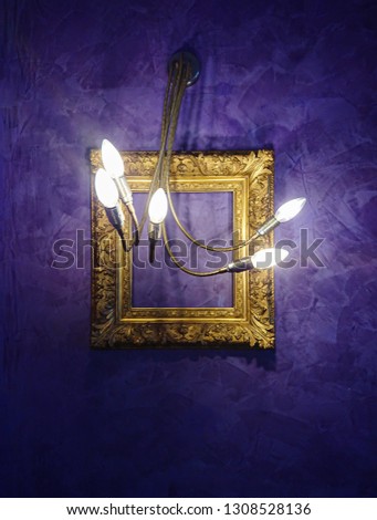 Modern design chandelier and golden picture antique frame in single composition on textured purple wall. Abstract decor interior