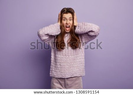 Young good looking woman screams in terror, opens mouth widely, covers ears, has irritated facial expression, wears oversized sweater, models over lilac background, doesnt want to hear yelling