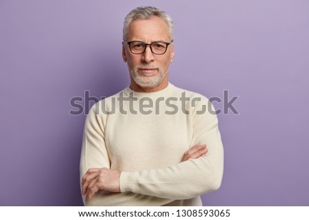 Headshot of confident mature European male pensioner keeps arms folded, wears optical glasses and white sweater, being sure in something, isolated over purple background. People and aging concept Royalty-Free Stock Photo #1308593065