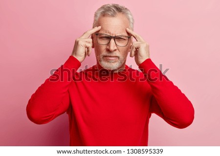 Concentrated retired man keeps fingers on temples, tries to recollect something in mind, has bad memory in old age, wears transparent spectacles and red sweater, models ober rosy studio wall Royalty-Free Stock Photo #1308595339