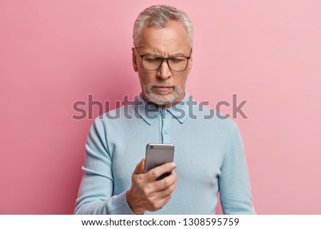 Serious mature man focused in screen of smartphone, connected to wireless internet, searches information, browses website, dressed in blue clothes, wears spectacles, isolated over rosy background