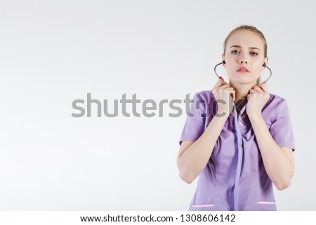 
Woman doctor blonde young light gray background studio day beautiful one purple lilac clothes stethoscope listen bad news sadness. Disease patient. Close up. Copy space left.