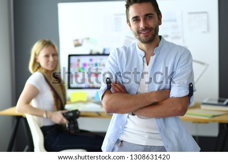 Portrait of young designer in front of laptop and computer while working. Assistant using her mobile at background.