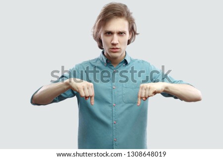 Here and right now. Portrait of serious handsome long haired blonde young man in blue casual shirt standing, looking at camera and pointing down. indoor studio shot, isolated on light grey background.