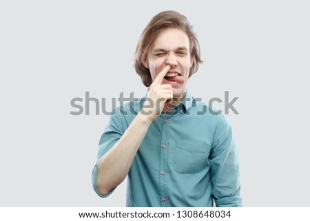 Portrait of funny crazy handsome long haired blonde young man in blue casual shirt standing drilling his nose and tongue out looking at camera. indoor studio shot, isolated on light grey background.