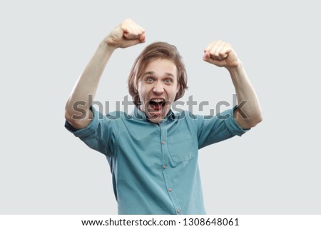 Portrait of amazed happy handsome long haired blonde young man in blue casual shirt standing screaming and celebrating his victory. indoor studio shot, isolated on light grey background.