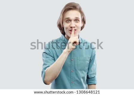 This is secret betwen us. Portrait of funny handsome long haired blonde young man in blue shirt standing looking at camera with silence sign. indoor studio shot, isolated on light grey background.
