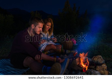 Man and woman baking sausages by the fire, drinking beer and relaxing by the campfire in the mountain top in the night. Romantic campfire concept.