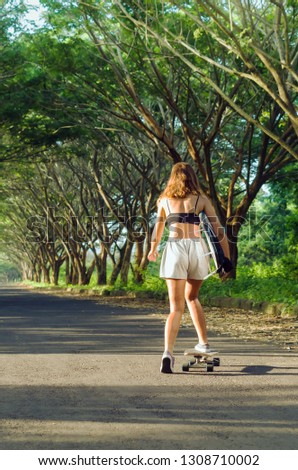 Girl with a surfboard riding on a skateboard towards the beach. Surfergirl in summer. 
