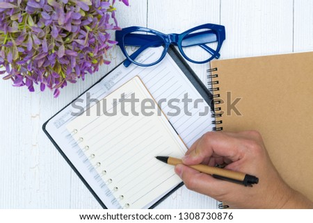 Man hand writing on blank page of notebook paper with pen on the wood, glasses and office supplies, top view.