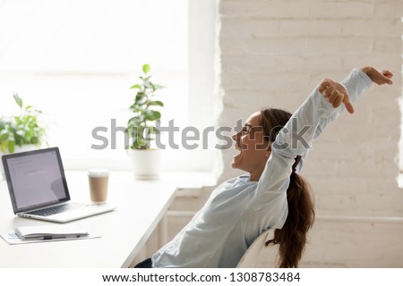 Cheerful mixed race woman sitting at workplace on chair bending stretching raising hands up, feels happy got a long-awaited post winning online lottery or accomplishing working day before vacation Royalty-Free Stock Photo #1308783484
