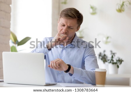 Exhausted or bored man sitting at desk in office looking at watch. Businessman yawning covering mouth with fist feels tired after busy hard working day. Chronic fatigue lack of energy or time concept
