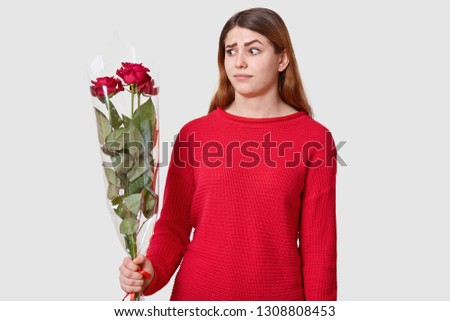 Indoor shot of dissatisfied woman raises eyebrows, looks in bewilderment, holds bouquet of flowers, doesnt expect to recieve roses from colleague, wears red jumper, has long straight dark hair