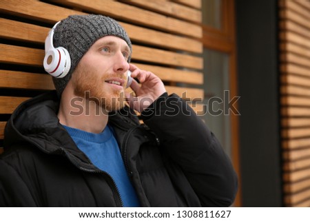 Young man listening to music with headphones near wooden wall. Space for text