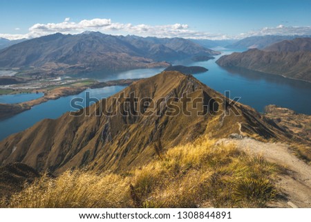 Picture of New Zealand’s beautiful Landscape from Mountain „Rois Peak“. The green/yellow mountain landscape with blue water makes out most of the picture.