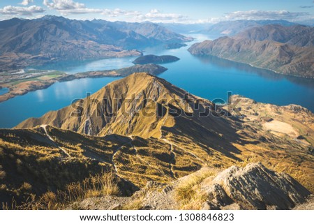 Picture of New Zealand’s beautiful Landscape from Mountain „Rois Peak“. The green/yellow mountain landscapes with hiking trails make up most of the picture. Blue lake in background.