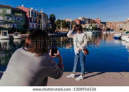two friends in the street taking pictures with mobile phone. Port background in a sunny day. Lifestyle outdoors. friendship concept