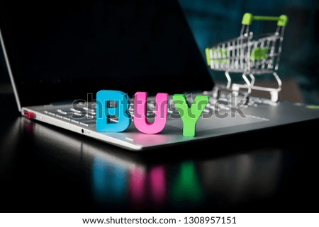 Creative shot of a wooden many-colored word BUY placed on laptop bottom at black screen and toy shopping trolley background. Concept of buying things in internet. Online shop promotion. E-business