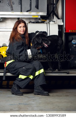 Image of young woman firefighter with black dog sitting on background of fire truck