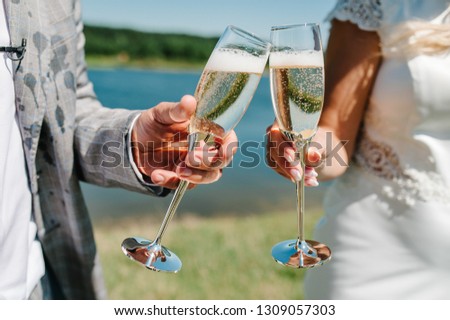 The bride and groom holds a glass of champagne and stand on nature at the wedding ceremony. Close up. Holiday. Look at the glasses. Toast.
