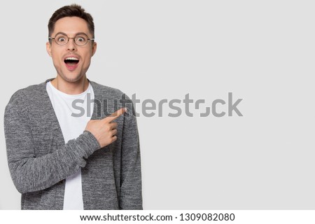 Amazed funny man in glasses pointing finger aside at copy space excited by unbelievable offer looking at camera, surprised male customer advertise product service isolated on studio blank background