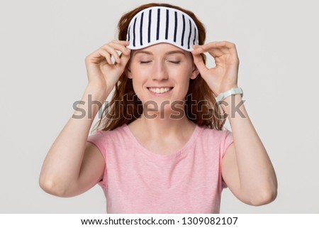 Smiling happy young redhead woman wearing striped sleeping eye mask feeling fresh after good night healthy sleep in the morning isolated on white grey studio background, comfortable sleepwear concept