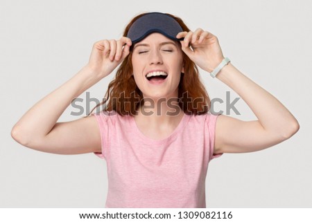 Happy funny red-haired woman laughing wearing grey sleeping eye mask, cheerful redhead lady enjoying comfortable sleepwear and good sleep isolated on white studio background, pajama party concept