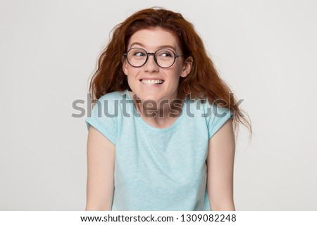 Cute shy awkward redhead teen girl in glasses biting lips feeling embarrassed, funny young red-haired woman student confused nervous before exam isolated on white grey blank studio background