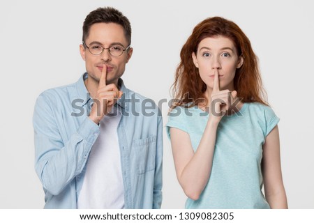 Young mute couple holding fingers on lips mouth to keep it quiet hush secret showing shh shush gesture for conspiracy silence looking at camera isolated on white grey blank studio background
