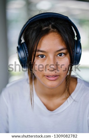 Close up portrait head shot of a young and attractive Chinese Asian girl. The millennial teenager is looking at the camera and smiling. She is wearing headphones as she enjoys listening to music. 