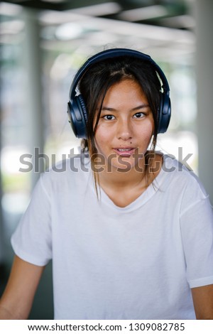 Close up portrait head shot of a young and attractive Chinese Asian girl. The millennial teenager is looking at the camera and smiling. She is wearing headphones as she enjoys listening to music. 