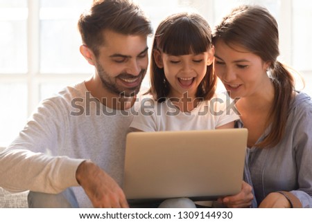 Cheerful happy mom dad and little kid daughter using laptop together at home, loving young family with child girl laughing looking at computer screen, enjoying shopping or watching cartoons online