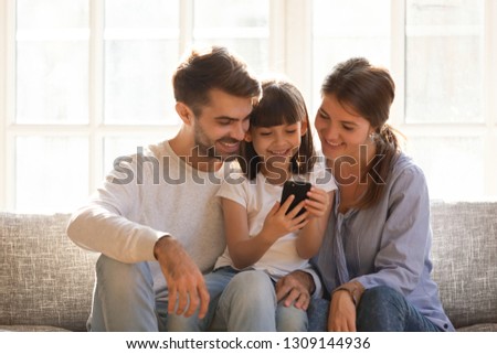 Little kid daughter holding cellphone enjoy using mobile app, happy family with child having fun with smartphone device make online call looking at phone screen, shop in application at home on couch