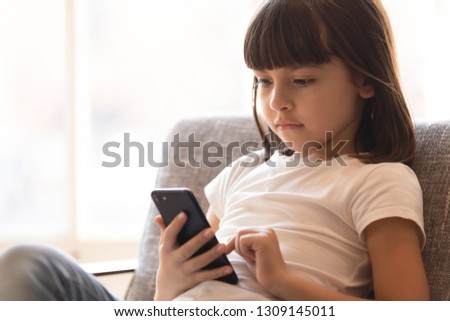 Curious cute preschool small girl using phone apps looking at cellphone device sitting on couch, little smart kid holding smartphone playing mobile game online alone at home, child and gadget concept