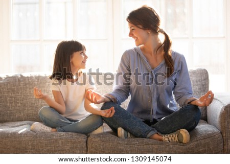 Mindful young mom teaching yoga happy cute funny child daughter sitting in lotus pose on couch together, smiling mother and little kid girl meditating relaxing at home, healthy family exercises Royalty-Free Stock Photo #1309145074