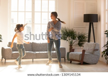 Happy young mom and cute little daughter jumping dancing in modern house living room, joyful mother with child girl having fun enjoy moving to music together at home, mum and kid funny activity
