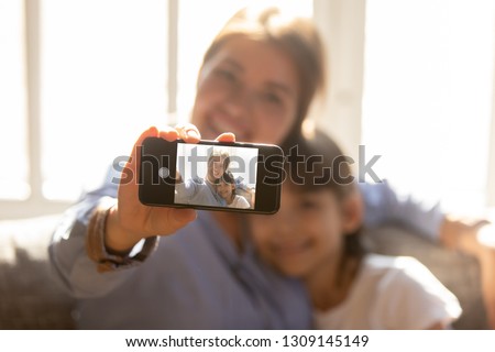 Happy mother embracing child daughter holding phone taking selfie, smiling mom and kid girl looking at smartphone camera making photo together, focus on gadget display with self portrait on screen