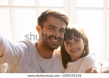 Dad and kid daughter with happy faces embracing posing taking selfie making photo or online call on phone, smiling father with child girl hugging looking at smartphone camera recording video at home
