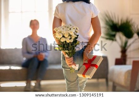 Rear view at little kid daughter holding flowers and gift box behind back congratulating happy young mom with mothers day or birthday, child girl hiding bouquet present making surprise to mum at home