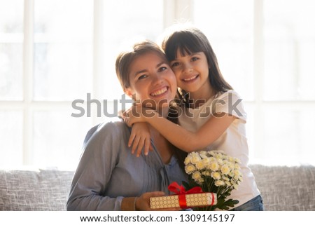 Happy little kid daughter embracing grateful mom holding flowers bouquet and gift box celebrate mothers day or birthday, cute child girl congratulating mum hugging looking at camera, family portrait