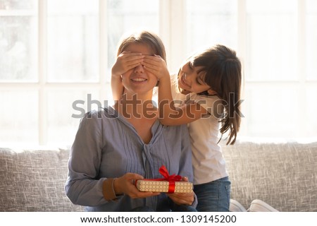 Happy little daughter covering mum eyes congratulating happy mom holding birthday present in gift box, smiling small kid girl making surprise to mommy on mothers day concept sitting on couch at home