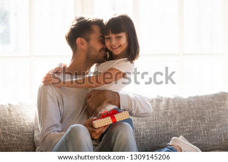 Grateful loving dad hugging little kid daughter thanking for gift on fathers day, happy daddy embracing smiling child girl with gratitude for birthday present holding box sitting on couch at home